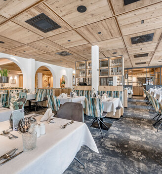 A spacious, elegant restaurant featuring a wooden ceiling, large windows, white tablecloths and meticulously set tables. Upholstered chairs and modern décor create an inviting and elegant atmosphere.