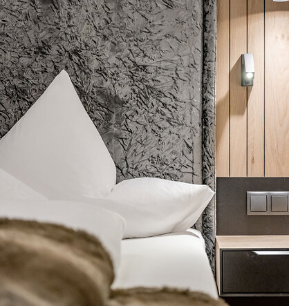 A modern hotel room with a neatly made bed, white pillows, a fluffy brown blanket, a wooden nightstand, power outlets and a wall-mounted reading lamp.