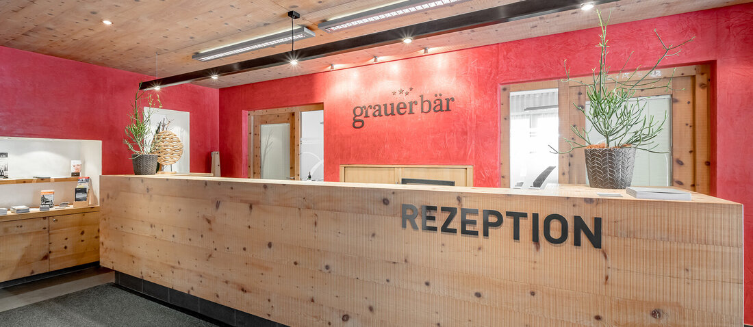 A modern hotel lobby featuring a wooden reception desk. The wall behind is red. Two potted plants stand on the counter, the area feels clean and welcoming.