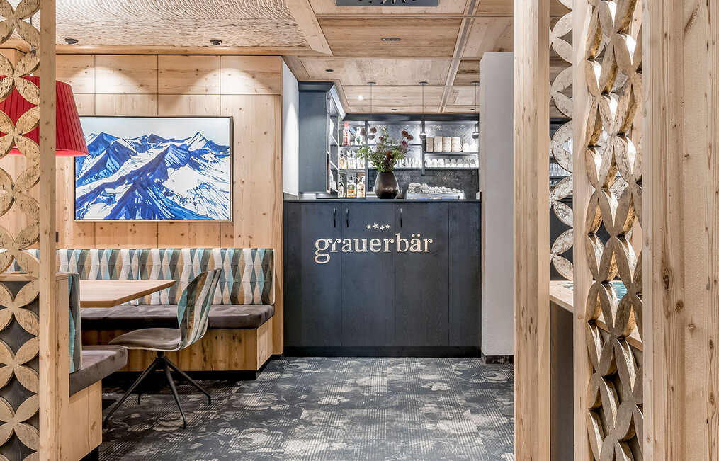 A modern restaurant interior featuring wood walls, patterned chairs, a mountain painting and a dark cabinet with the inscription "Grauer Bär." Decorative wood panels frame the cozy dining area.
