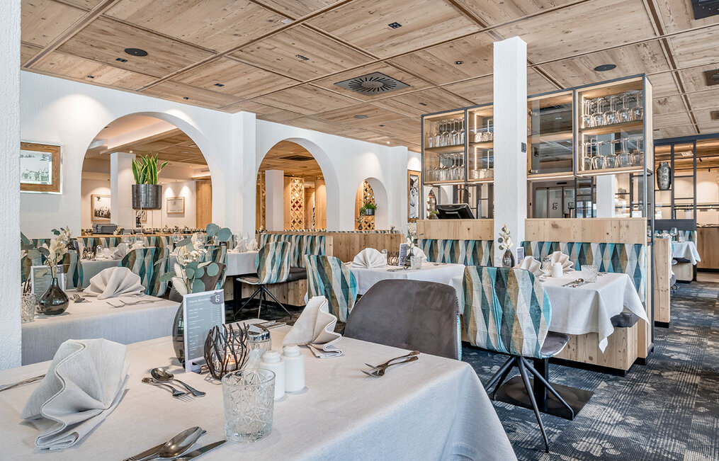 A bright, modern restaurant interior featuring wooden ceilings, tables set with white linens, patterned booth seating and ornamental plants. Glassware and tableware are neatly arranged, creating an inviting atmosphere for dining.