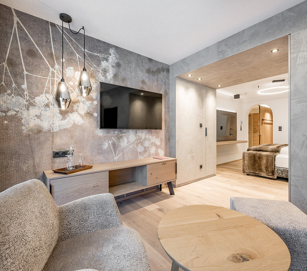 Modern hotel room with textured gray walls, a wall-mounted TV, a wooden sideboard, two upholstered armchairs and a round coffee table. Pendant lights hang from the ceiling, a bedroom is behind an open door.