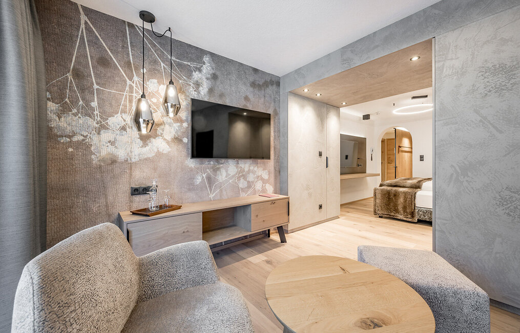 Modern hotel room with textured gray walls, a wall-mounted TV, a wooden sideboard, two upholstered armchairs and a round coffee table. Pendant lights hang from the ceiling, a bedroom is behind an open door.