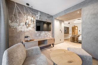 Modern hotel room with textured gray walls, a wall-mounted TV, a wooden sideboard, two upholstered armchairs and a round coffee table. Pendant lights hang from the ceiling, a bedroom is behind an open door.