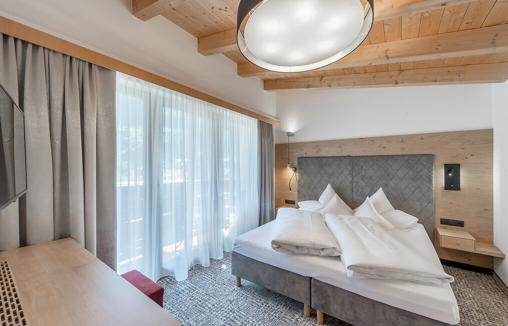Modern hotel room with a large bed, a gray headboard, white bedding, a desk, a wall-mounted TV, a large window with sheer curtains, a wooden ceiling and a round ceiling light.