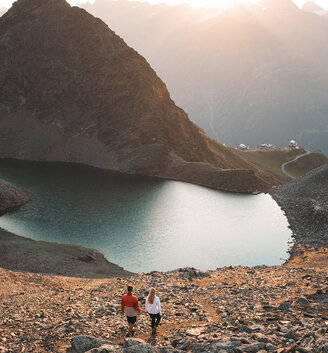 Two people hike down a rocky slope toward a mountain lake at sunset with mountains and a winding path in the background.
