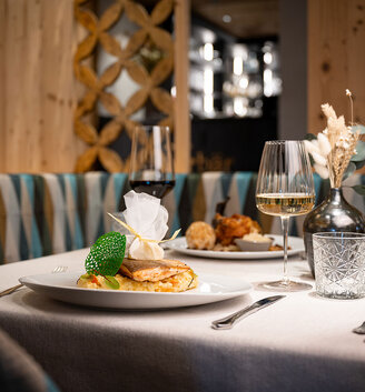 A restaurant table set for two, featuring plates filled with gourmet dishes, glasses of red and white wine, a small vase of dried flowers and elegant table décor - all within a cozy, modern setting.