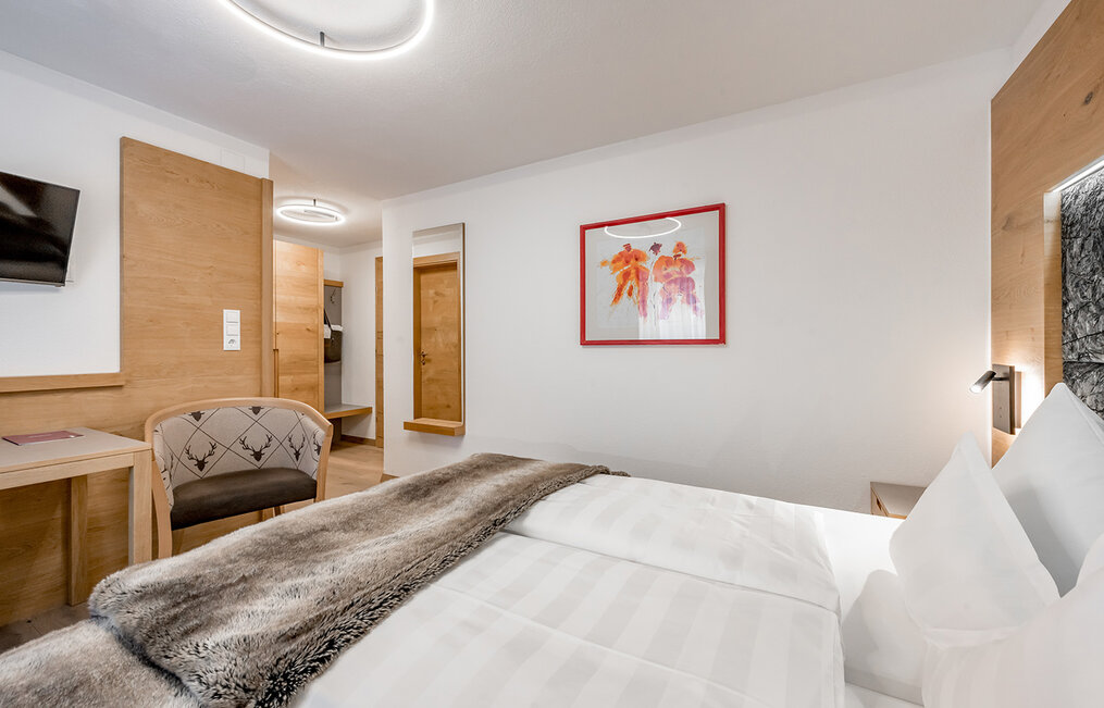 A modern hotel room featuring a neatly made bed, a fur throw, wooden furniture, a chair, a desk, a wall-mounted TV and a colorful, framed artwork above the bed. The room is bright and features minimalist décor.