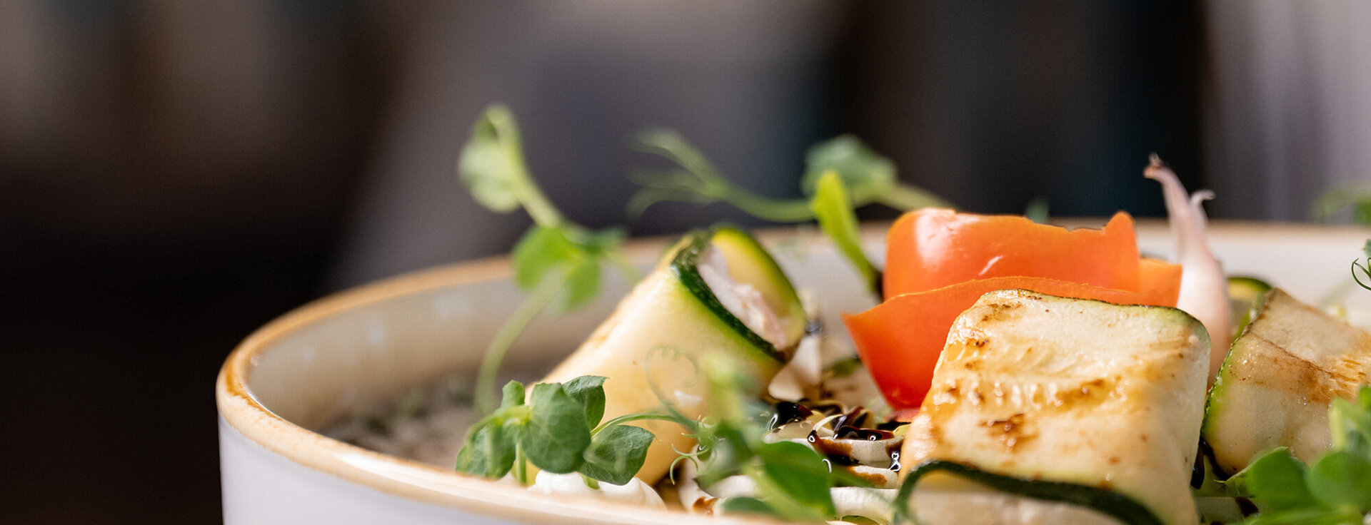 A close-up of a gourmet dish featuring grilled zucchini rolls, microgreens and a tomato slice, artfully arranged on a white plate against a blurred background.