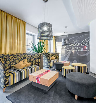 Modern lounge featuring a patterned sofa, yellow curtains, round floor cushions, wooden tables, pendant lights and decorative plants. The walls are adorned with lettering. A bright, inviting space offering a contemporary ambiance.