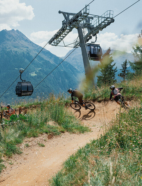 Three cyclists are riding downhill along a dirt path through grassy terrain. A mountain gondola and an alpine landscape are in the background while wildflowers bloom in the foreground.