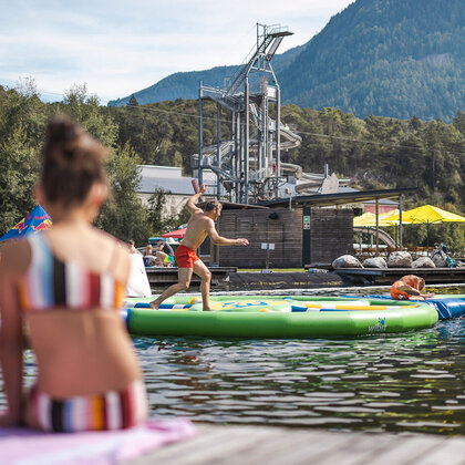 Two people are sitting on a jetty, watching people play on a colorful, floating obstacle course in a swimming lake while trees, mountains and water slides are in the background.