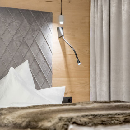 A neatly made bed with white pillows and a duvet, a grey upholstered headboard, an adjustable reading lamp and a hanging pendant light, alongside beige curtains and a wooden accent wall.