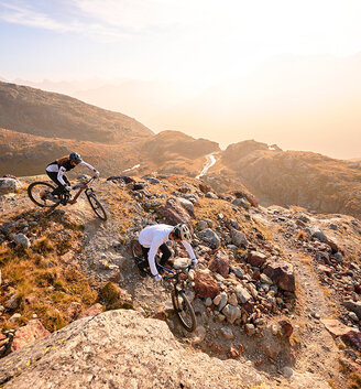 Two mountain bikers ride down a rocky path on a rugged slope at sunset, surrounded by mountains and a glowing sky in the background.
