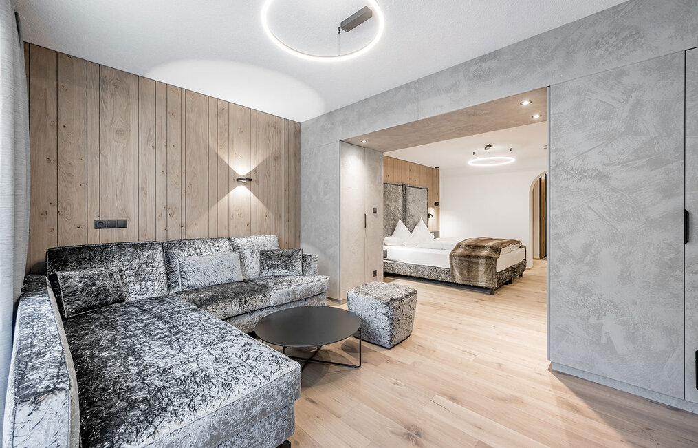 A modern hotel suite featuring a sophisticated silver sofa and a round coffee table in the foreground. In the background, a double bed with white bedding and a fur throw stands beneath soft ceiling lighting.