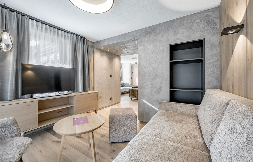 Modern living room with a beige sofa, a round wooden coffee table, a TV on a wooden stand, open shelving and a large window with sheer curtains. Neutral tones and minimalist decor.