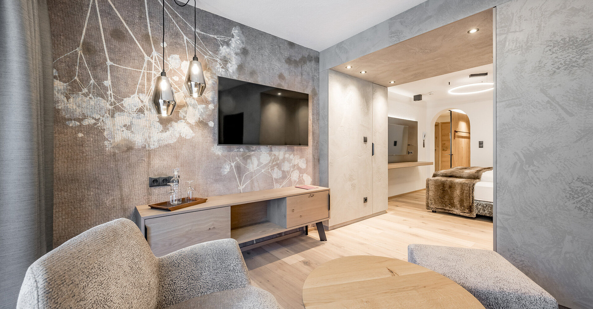 Modern hotel room with textured walls, a flat-screen TV, two upholstered armchairs, a round wooden table, pendant lights and a view of a sleeping area with a large bed.