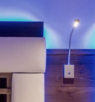 A modern reading lamp with a flexible arm is attached to a wooden headboard, which is illuminated by soft blue LED accent lighting. Upholstered panels are mounted on both sides of the headboard.