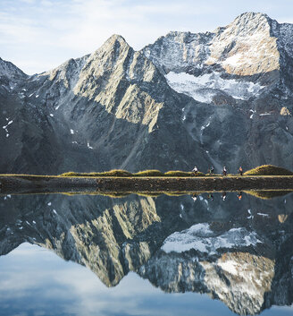 Snow-capped mountains are reflected in a calm lake with a grassy shore separating the water from the peaks under a partly cloudy sky.