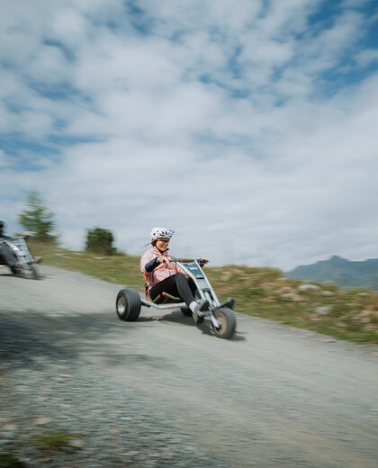 Two people are riding tricycles down a gravel path on a hillside. Both are wearing helmets. In the background, there is a cloudy sky with distant mountains and a green landscape, conveying a sense of movement and speed.