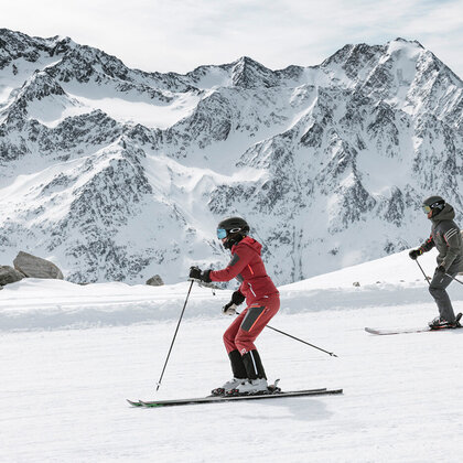 Two people skiing on a snowy slope, with rocky, snow-capped mountains in the background under a partly cloudy sky.