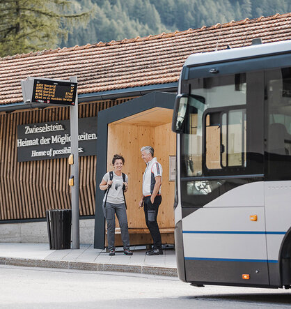 Zwei Personen stehen an einer Bushaltestelle und unterhalten sich, wobei ein Bus die Sicht teilweise versperrt. Auf dem Schild der Bushaltestelle sind die Fahrpläne angegeben. Hinter ihnen befindet sich ein Holzgebäude mit Ziegeldach.