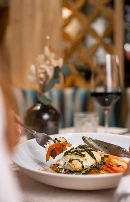 Eine Person schneidet ein Gericht mit Tomaten, Mozzarella und Pesto auf einem weißen Teller an, im Hintergrund stehen ein Glas Rotwein und eine Vase mit Blumen.