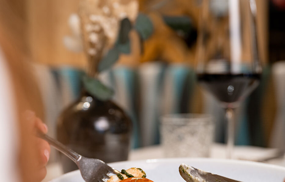 A person is cutting into a dish with tomatoes, mozzarella and pesto on a white plate. In the background, there is a glass of red wine and a vase of flowers.
