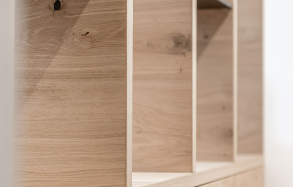 A light-colored wooden shelving unit with vertical and horizontal dividers and base cabinets featuring metal handles, positioned against a white wall. The shelves are empty and feature a minimalist design.