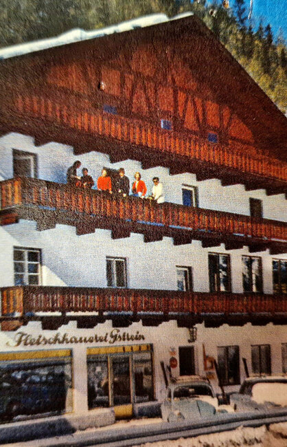 Ein Gebäude mit Holzbalkonen, Menschen, die draußen und auf dem Balkon stehen, verschneiter Boden, geparkte Autos, Kiefern und schneebedeckte Berge im Hintergrund.