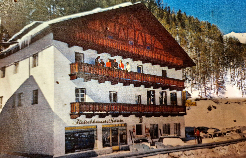 A building with wooden balconies, people standing outside and on the balcony, snow-covered ground, parked cars, pine trees and snow-capped mountains in the background.