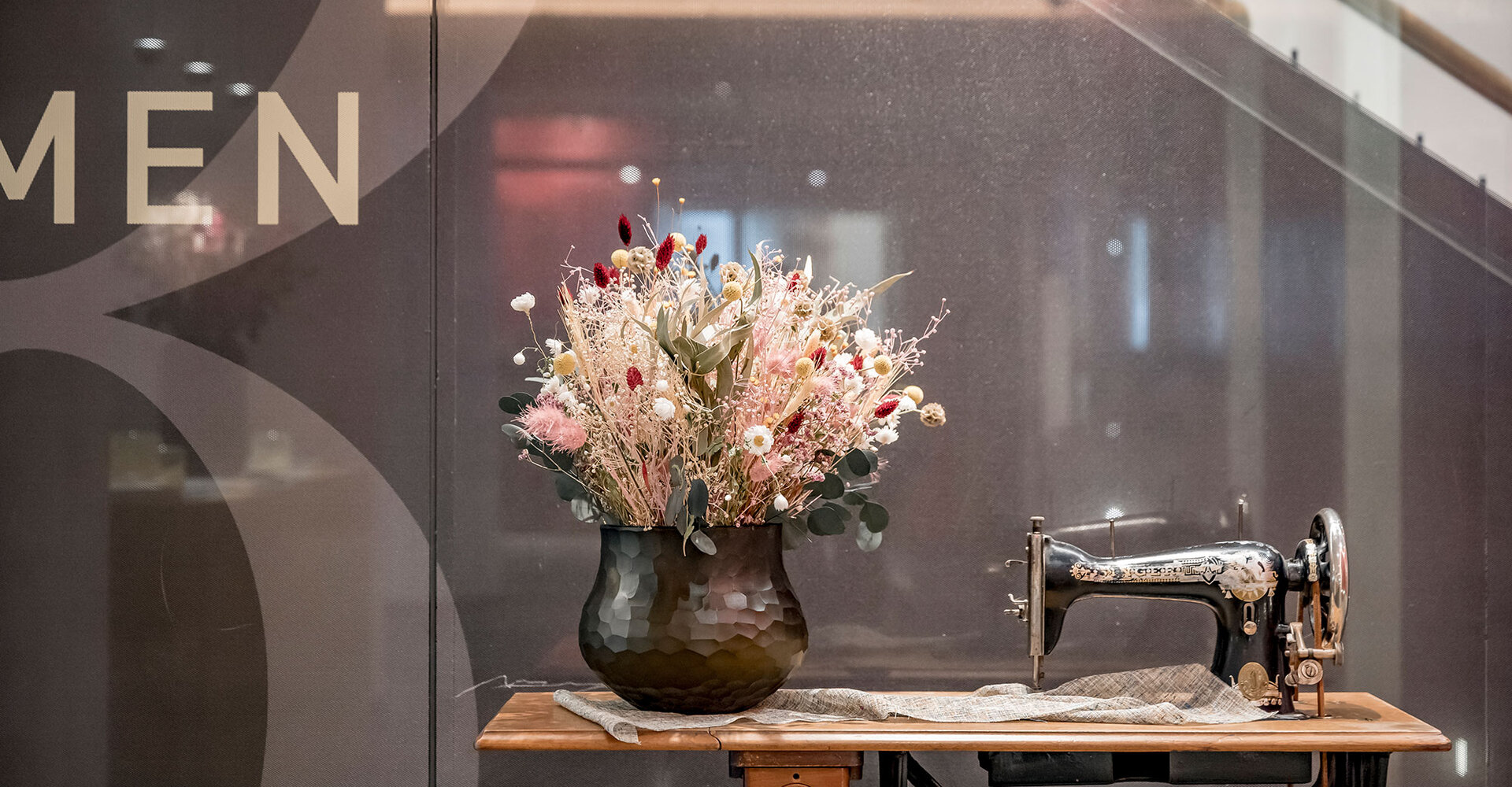 An old sewing machine is placed on a wooden table next to a black vase filled with colorful dried flowers. In the background there is a glass wall with some lettering partially discernible on the left side.