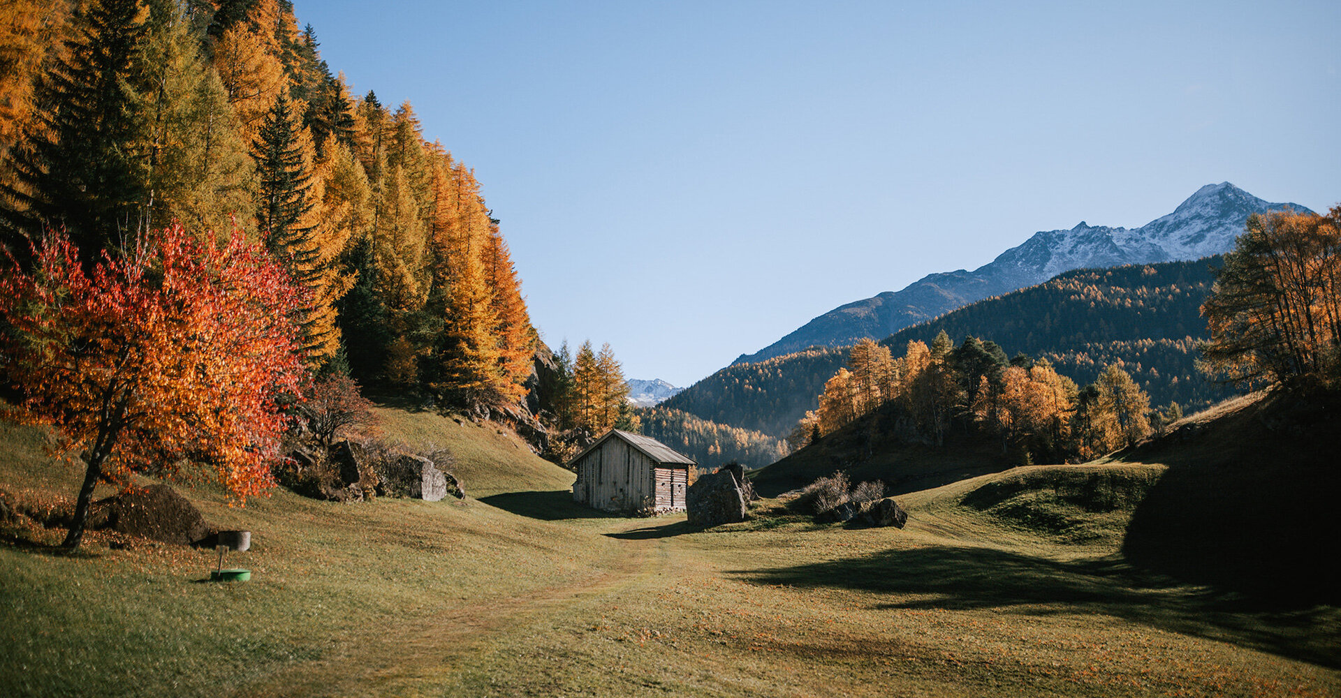 In a grassy valley, surrounded by trees colored in autumn, stands a small hut while in the background, mountains rise against a bright blue sky.