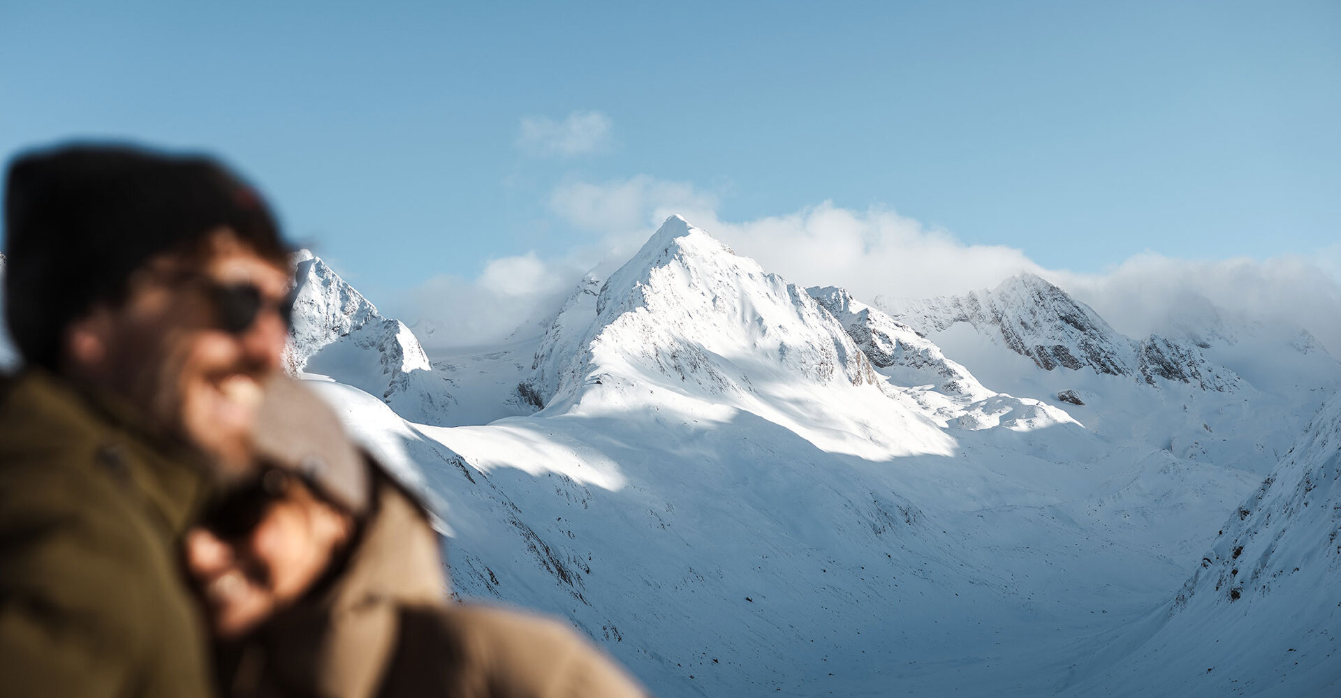 In the foreground are two people in winter clothing, depicted out of focus, while in the background there are snow-covered mountain peaks against a blue sky.