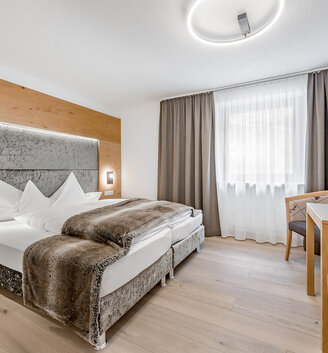 Modern hotel room featuring a double bed, a faux-fur throw, wooden furniture, a desk with a chair, a wall-mounted TV and a large window with curtains and blackout drapes. The room is decorated in neutral tones and features modern lighting.