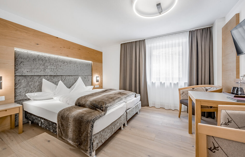 Modern hotel room featuring a double bed, a faux-fur throw, wooden furniture, a desk with a chair, a wall-mounted TV and a large window with curtains and blackout drapes. The room is decorated in neutral tones and features modern lighting.