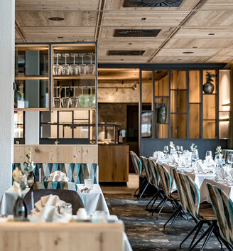 A modern restaurant area featuring a wooden ceiling, large windows and tables set with white tablecloths, glassware and folded napkins. Empty chairs are neatly arranged, additional glassware is displayed on shelves.