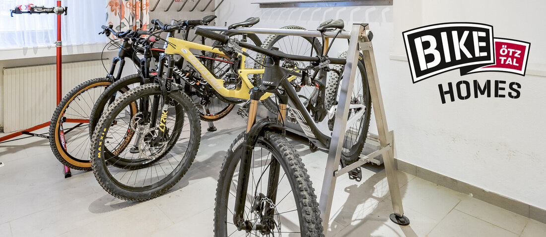 Several mountain bikes stand in a bicycle storage room equipped with a bike rack, a repair stand, wall hooks and a nature photograph on the wall. Light streams in through a window with patterned curtains.