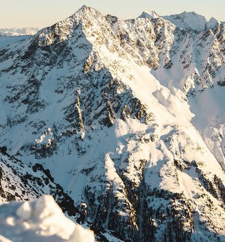 The snow-capped mountain peaks are illuminated by golden sunlight under a clear sky, with deep shadows accentuating the rugged structure of the landscape.