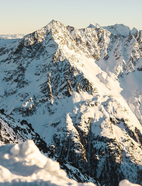 The snow-capped mountain peaks are illuminated by golden sunlight under a clear sky, with deep shadows accentuating the rugged structure of the landscape.