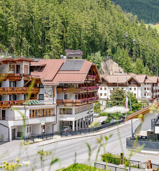 The hotel building is surrounded by dense green forest and mountains. A road runs through the foreground, flanked by blooming flowers and abundant greenery. It is a bright, sunny day.