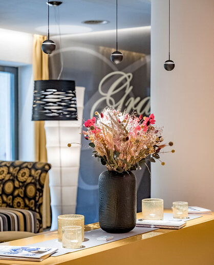 A modern reception desk featuring a black vase filled with pink and white flowers, surrounded by candles. Pendant lights hang above, patterned furniture in the background.