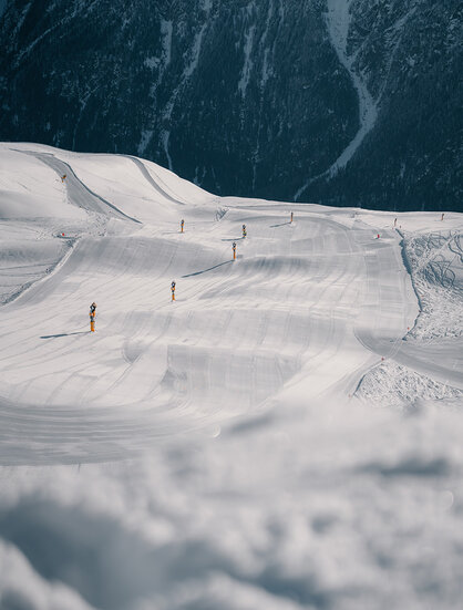 A snow-covered ski slope with several yellow markers winds through a mountainous landscape.
