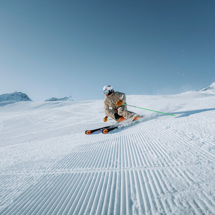 A skier in light brown ski wear and a white helmet speeds down a groomed, snow-covered slope under a brilliant blue sky, with mountains in the background.