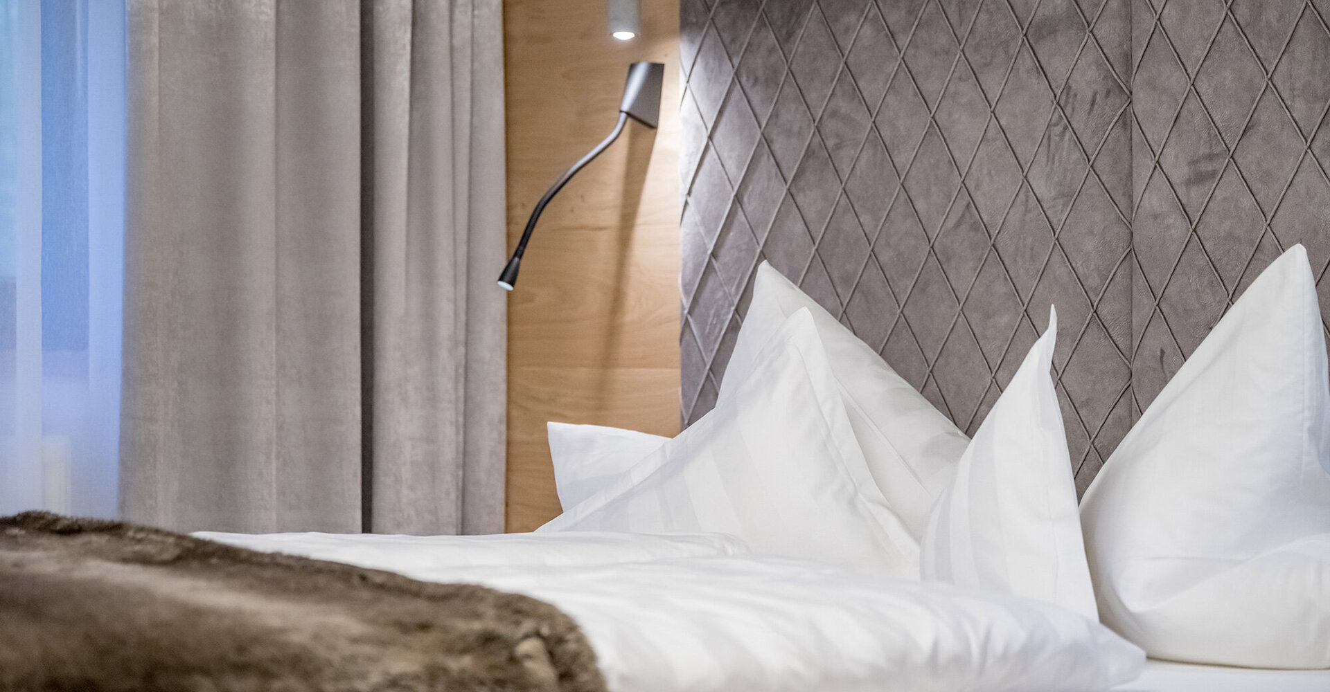 Ein modernes Hotelbett mit einem grauen Kopfteil im Rautenmuster, weißen Kissen, einer weißen Bettdecke, einer Fell-Decke, grauen Vorhängen und einer an der Holzwand befestigten Leselampe.