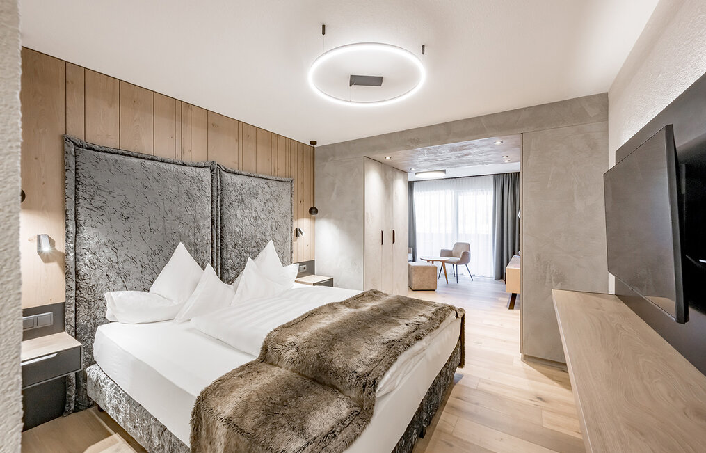 Modern hotel room featuring a double bed, a fluffy blanket, wood paneling, a large headboard, a TV, a desk and a seating area by the window with sheer curtains. The room is bright and decorated in neutral, natural tones.