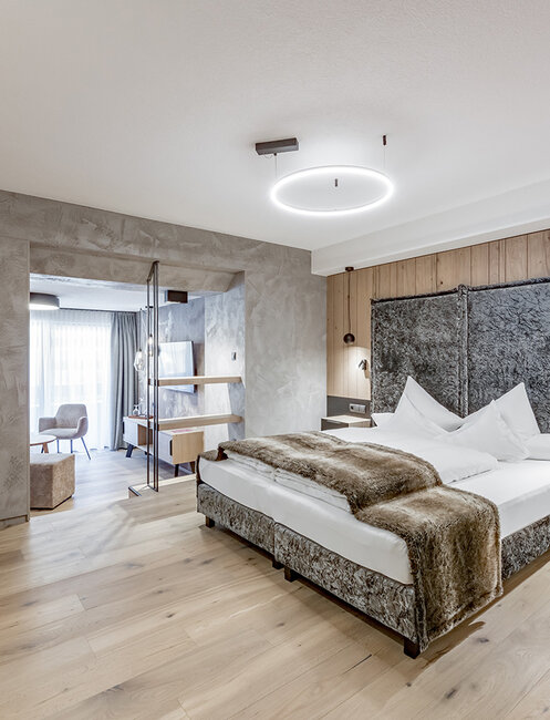 Modern hotel room featuring wooden floors, a double bed, a wall-mounted TV and a seating area with chairs next to a large window that lets in plenty of natural light. The décor is minimalist and features neutral tones.
