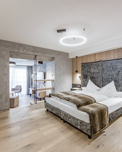 Modern hotel room featuring wooden floors, a double bed, a wall-mounted TV and a seating area with chairs next to a large window that lets in plenty of natural light. The décor is minimalist and features neutral tones.