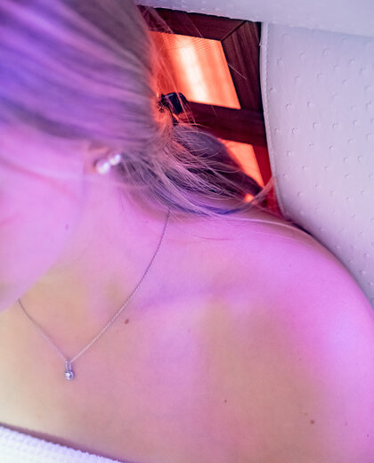Eine Frau mit blonden Haaren, die eine Halskette und ein Handtuch trägt, sitzt unter einem violett schimmernden Licht, auf einem Infrarot-Stuhl. Ihr Gesicht ist teilweise außerhalb des Bildausschnitts.