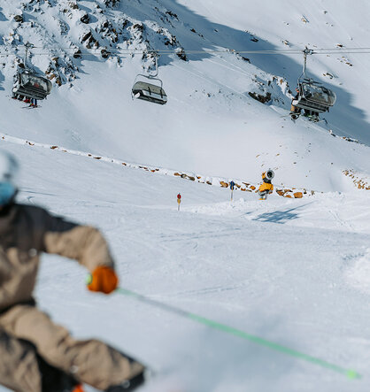 A skier in brown gear speeds down the slope, leaving a blur in the foreground. In the background, snow-capped mountains, ski lifts and other skiers are under a clear sky.